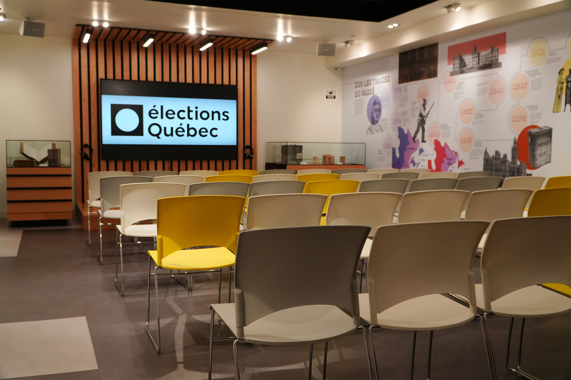 Salle de conférence avec chaises grises et jaunes, écran affichant Élections Québec, mur décoré d’images variées.