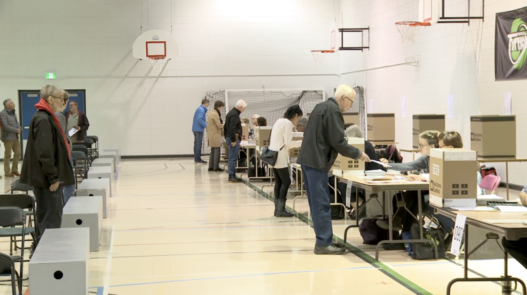 Plan large d’un lieu de vote dans un gymnase. À gauche de la photo, des gens patientent pour aller à leur bureau de vote et, à droite, cinq électrices et électeurs se tiennent debout devant des tables de vote afin de recevoir leur bulletin de vote.
