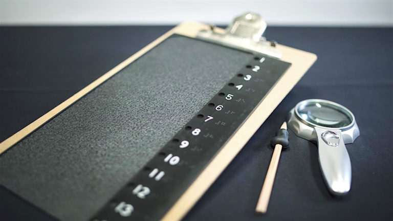 Vue rapprochée de quatre outils offerts pour aider les gens à voter : une tablette avec pince en haut pour maintenir le bulletin de vote, une règle numérotée de 1 à 14 en braille, un crayon avec embout en caoutchouc permettant une meilleure prise et une loupe.